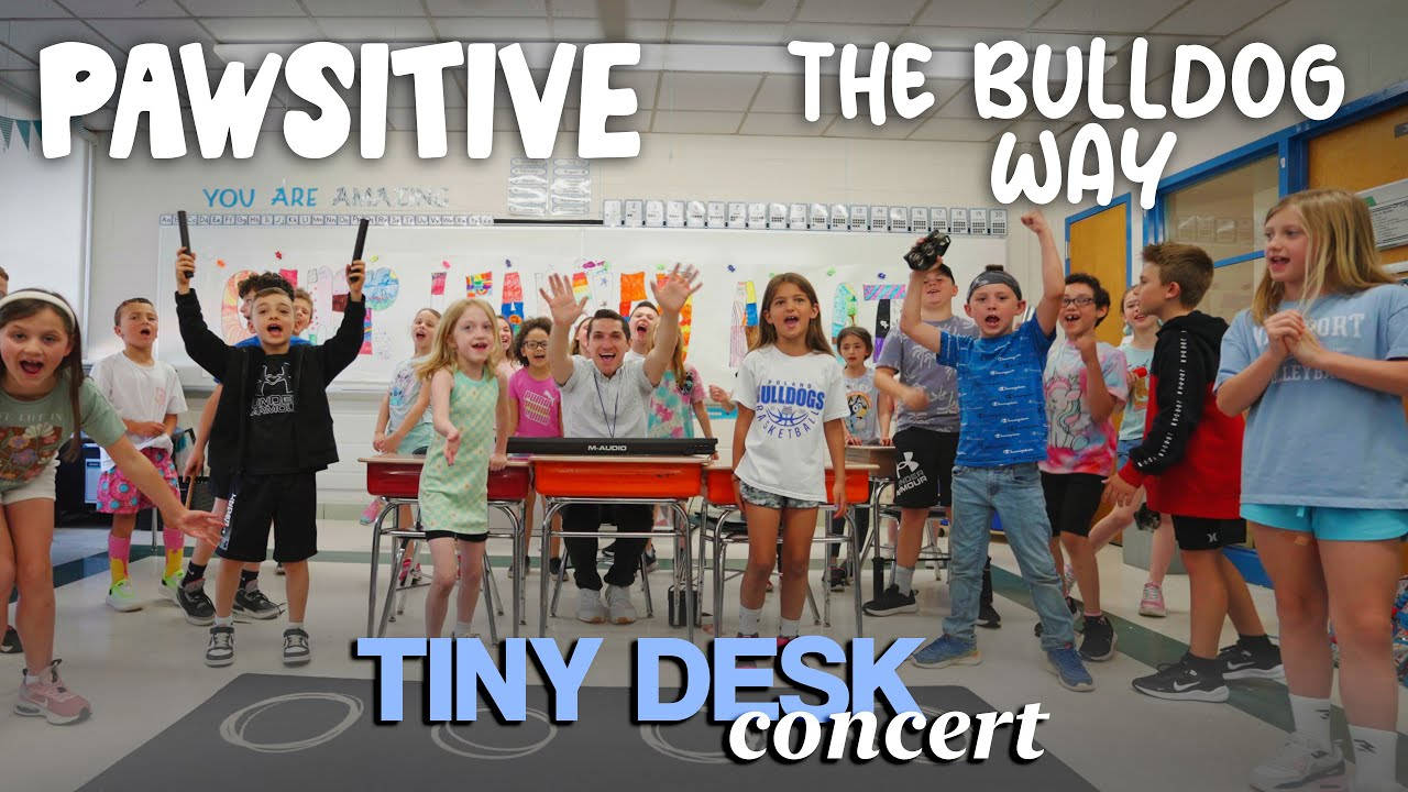 A joyful class of elementary students surrounds a keyboard, singing and dancing during a Tiny Desk-style concert promoting positivity the Bu